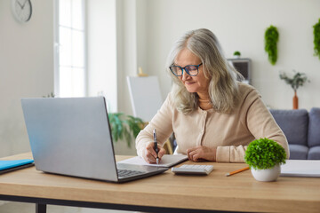 Senior woman sitting at desk, home, older writing in notebook using laptop, calculate budget, motivated learning or working digitally, active seniors technology, online engagement busy study 