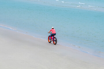 Obraz premium Carefree woman in sports fitness clothes riding Fat bike also called fatbike or fat-tire bike in summer driving on beach. Active healthy lifestyle. Enjoy and fun with bicycle on sand at coastal.