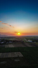 The golden sun sets over the ocean, casting a warm, beautiful glow across a vast patchwork of rural farmland and a coastal village below.