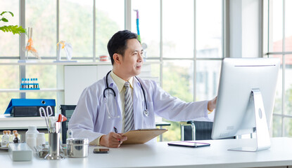 Asian male doctor in white coat with stethoscope, sitting reading paperwork for results at desk in modern hospital medical clinic office, research professional healthcare insurance wellness technology