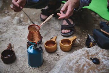 A person brews coffee outdoors using a copper pot on a portable stove, surrounded by wooden cups, depicting a serene and rustic camping experience.