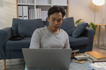 A young man is working from home, sitting on the floor with his laptop and smartphone, surrounded...