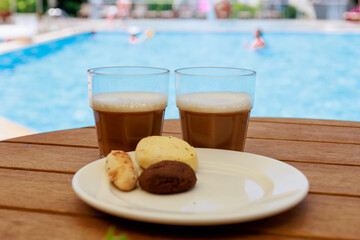 Glasses of iced coffee and cookies near the pool on blurred nature background. Vacation, resort, summer, travel concept.