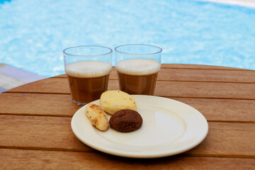 Glasses of iced coffee and cookies near the pool on blurred nature background. Vacation, resort, summer, travel concept.