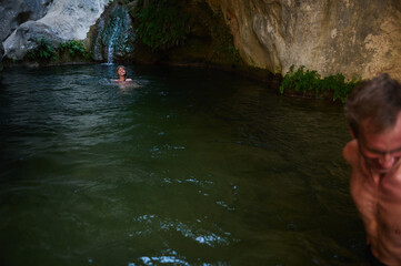 Two individuals enjoy swimming in a serene natural pool surrounded by rocks, moss, and greenery with a waterfall adding a tranquil ambiance.