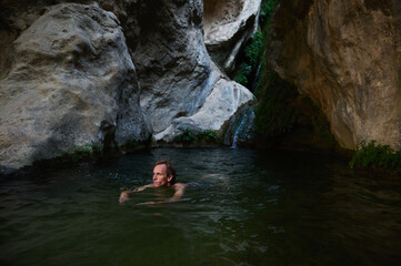 A man enjoying a refreshing swim in a serene rock pool nestled amidst rugged cliffs, with greenery and a small waterfall creating a peaceful atmosphere.