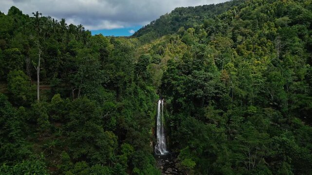 A stunning aerial drone shot of Gitgit Waterfall in Bali, flowing in full view, surrounded by tropical trees and vibrant green foliage, with clouds and blue sky visible above.