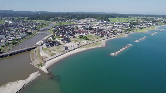 Aerial drone view of a coastal town in Noto Peninsula Suzu, Japan. Blue sea, breakwaters, rivers, and a mix of traditional and modern homes highlight this peaceful regional landscape on a sunny day.