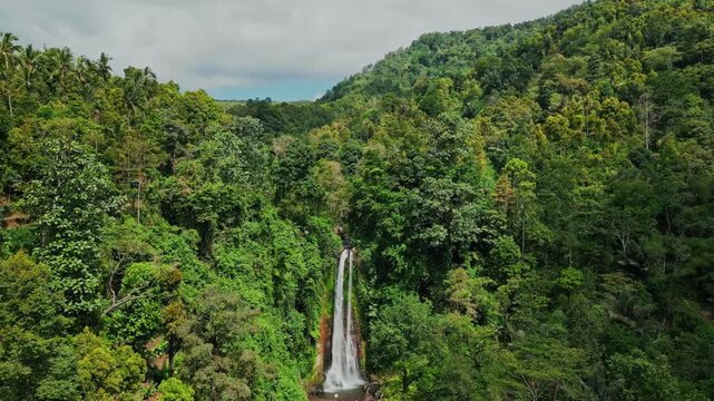 A stunning aerial drone shot of Gitgit Waterfall in Bali, flowing in full view, surrounded by tropical trees and vibrant green foliage, with clouds and blue sky visible above.