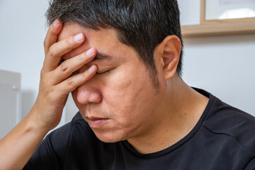 Fototapeta premium Middle-aged Asian man clutching his head, seated after long hours of work, experiencing headache and dizziness—ideal for medical ads on fatigue relief, neurology, wellness care.