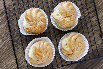 Four freshly baked Kue Sus (Choux Pastries or Cream Puffs) are neatly arranged on a black wire cooling rack. Each puff is perfectly golden-brown and filled with a visible, creamy filling.