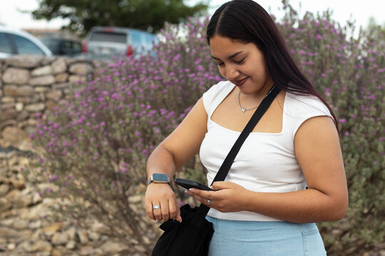 Young woman checking time on smartwatch while using smartphone outdoors