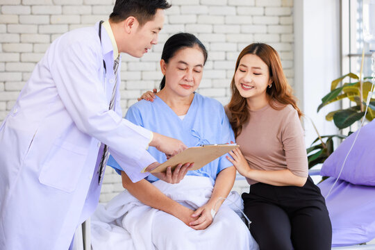 Asian man professional doctor visiting ward talking giving report good news to smiling and happy family and senior woman patient sitting on hospital bed with lovely daughter showing healthcare support