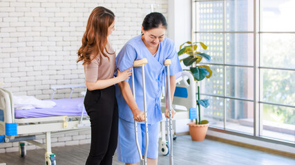 Fototapeta premium Asian young woman teaching using crutches walks beside senior female patient sitting on hospital bed medical recovery room caregiver specialist wellness healthcare treatment rehabilitation support.