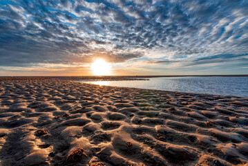 Sonnenuntergang &uuml;ber dem Watt bei Utersum auf der Nordseeinsel F&ouml;hr mit der Sonne am Horizont unter Sch&auml;fchenwolken, und einem Priel mit Strand mit Wellenmuster