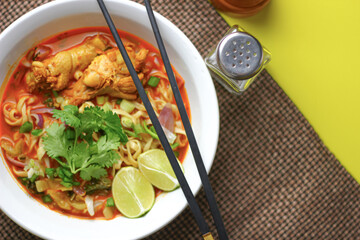 The image shows a bowl of Khao Soi, a traditional Thai noodle curry with chicken, crispy noodles, fresh cilantro, lime wedges, and pickled greens. The dish is beautifully arranged and vibrant.