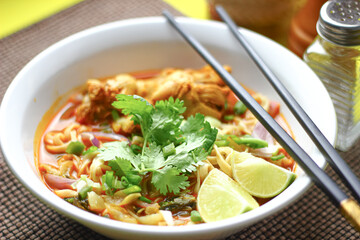 The image shows a bowl of Khao Soi, a traditional Thai noodle curry with chicken, crispy noodles, fresh cilantro, lime wedges, and pickled greens. The dish is beautifully arranged and vibrant.