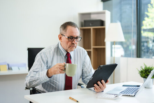 Senior businessman in formal attire using tablet and drinking coffee at modern office desk, working on finance, planning, or digital consulting tasks.