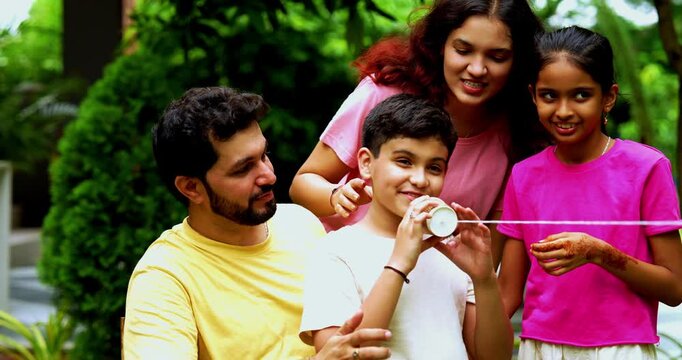 Indian family playing with string telephone in home garden, enjoying creative outdoor activity, spending quality fun time together with kids, parents, siblings, smiling and bonding joyfully