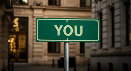 Obraz premium A green street sign with the word YOU in white letters against a blurred city background.