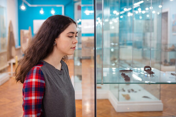 Young Caucasian woman visiting museum and viewing ancient pottery behind glass, concept of education and cultural appreciation during International Museum Day. Side view