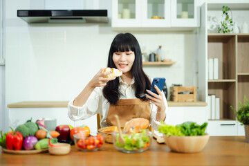 Smiling Asian woman enjoys a croissant and coffee in a modern kitchen with fresh vegetables. A...