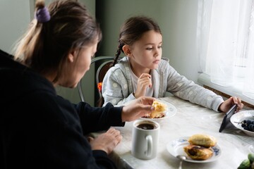 Girl with adult, enjoying a meal and looking at the phone by the window, light color, morning. Eating together at the table.