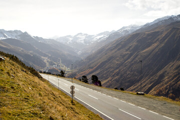 A winding asphalt road cuts through the rugged terrain of the Timmelsjoch Pass. Snow glistens on distant summits. Distant layers of mountains fade into a hazy horizon.