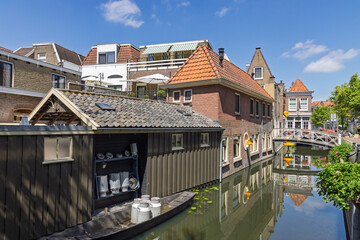 Fototapeta premium Traditional Dutch houses and a rustic wooden shed sit along a serene canal in Gouda, with a small boat and cheese molds.