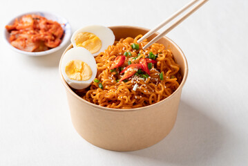 Spicy Korean dried instant noodles with boiled egg, sliced scallions, sesame seed and chili in kraft paper bowl and kimchi cabbage on white background