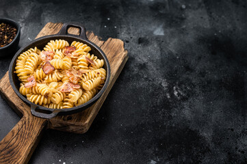 Fusilli carbonara pasta topped with crispy bacon bits and parmesan  cheese  in a skillet. Black background. Top view