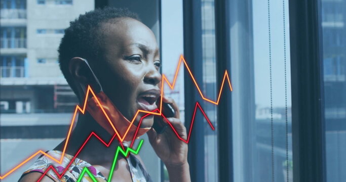 Speaking businesswoman holding smartphone at ear by office window, displaying colorful line graphs - Powered by Adobe