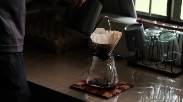 Medium static shot of Gooseneck kettle pouring hot water into V60 dripper and filter paper for coffee pour-over brew over glass carafe during morning.