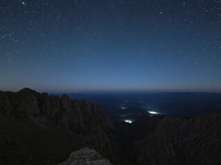 A night view from a mountain peak showing a dark landscape and a starry sky above the horizon