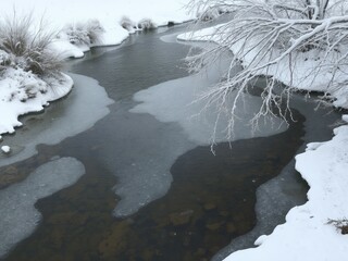A winter scene showing a partially frozen river with snow covered banks and tree branches