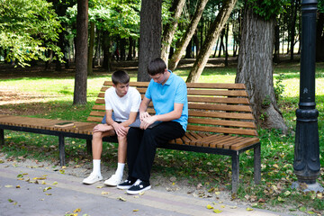 Fototapeta premium Two young men sit on a park bench, one showing something on a tablet. Lush greenery surrounds them in this outdoor scene. Casual day.