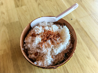 A woven bowl holds a generous serving of fluffy white rice, topped with crispy fried shallots, accompanied by a white plastic serving spoon.