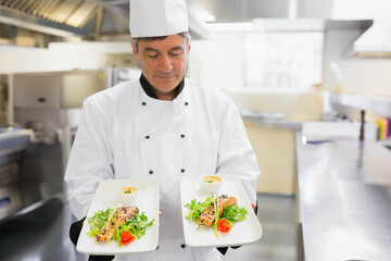 Senior male chef wearing chef coat presenting two seared fish plates with sauce, greens in kitchen