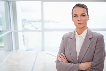 Middle-aged woman standing crossing arms in office with polished floor and windows, copy space