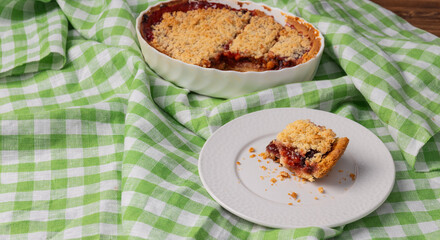 traditional plum crumble pie in ceramic baking dish with one slice served on white plate over green checkered cloth concept of homemade baking, food blog, rustic dessert