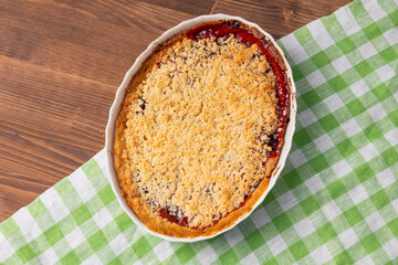 whole plum crumble pie in ceramic baking dish on wooden table with green checkered cloth. top view of homemade fruit dessert. concept of home baking, seasonal treats, food blog
