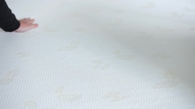A hand pressing gently on a white mattress featuring embossed leaf patterns and subtle textured fabric. Close-up highlighting surface softness and fabric responsiveness to touch