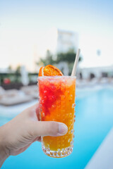 Hand holding a refreshing tropical cocktail with a dried orange slice and paper straw, poolside on a sunny day, creating a vibrant and inviting summer scene