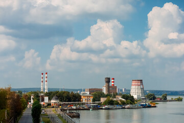 Naklejka premium On the river bank there is a thermal power plant with tall chimneys, surrounded by greenery. The sky is covered with fluffy clouds, creating a calm and picturesque view.