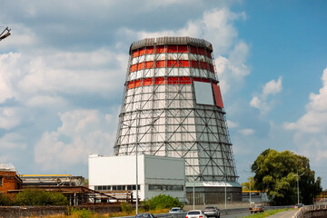 A tall cooling tower of a thermal power plant surrounded by greenery and roads. The sky is clear, with clouds creating a contrast with the industrial object.