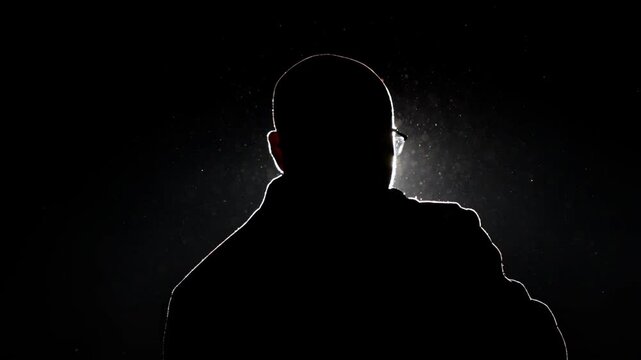 A man with glasses sings into a microphone in the dark against a backlight. The outline of dust and light in the shadow. Performance on stage, slow mo