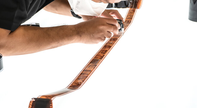 A man closely examining a developed color film strip under magnification on a white background.
