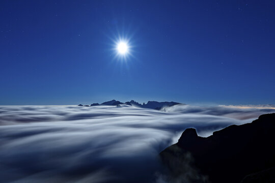 View of a celestial dance unfolds as moonlight bathes ethereal clouds, hugging rugged peaks in a tranquil embrace, creating a dreamscape, Madeira, Portugal.