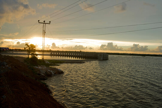 A peaceful lake scene at sunset with golden light reflecting off the water, dramatic clouds, and power lines crossing above.