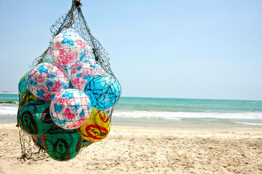 Colorful balls hang on a beachside net with waves, sand, and ocean in the background.